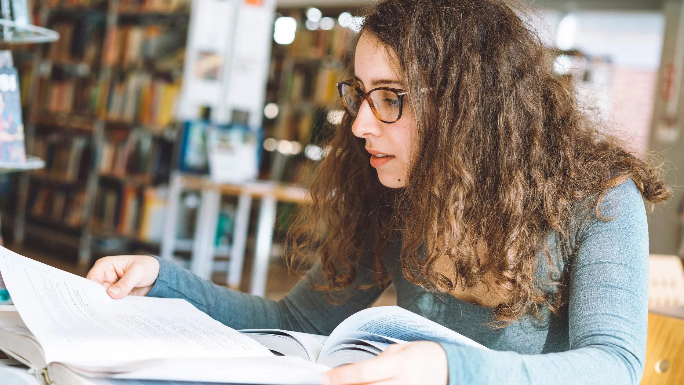 A young woman with curly hair and glasses studies for USMLE Step 1 and Step 2 in a library, intently reading a textbook.